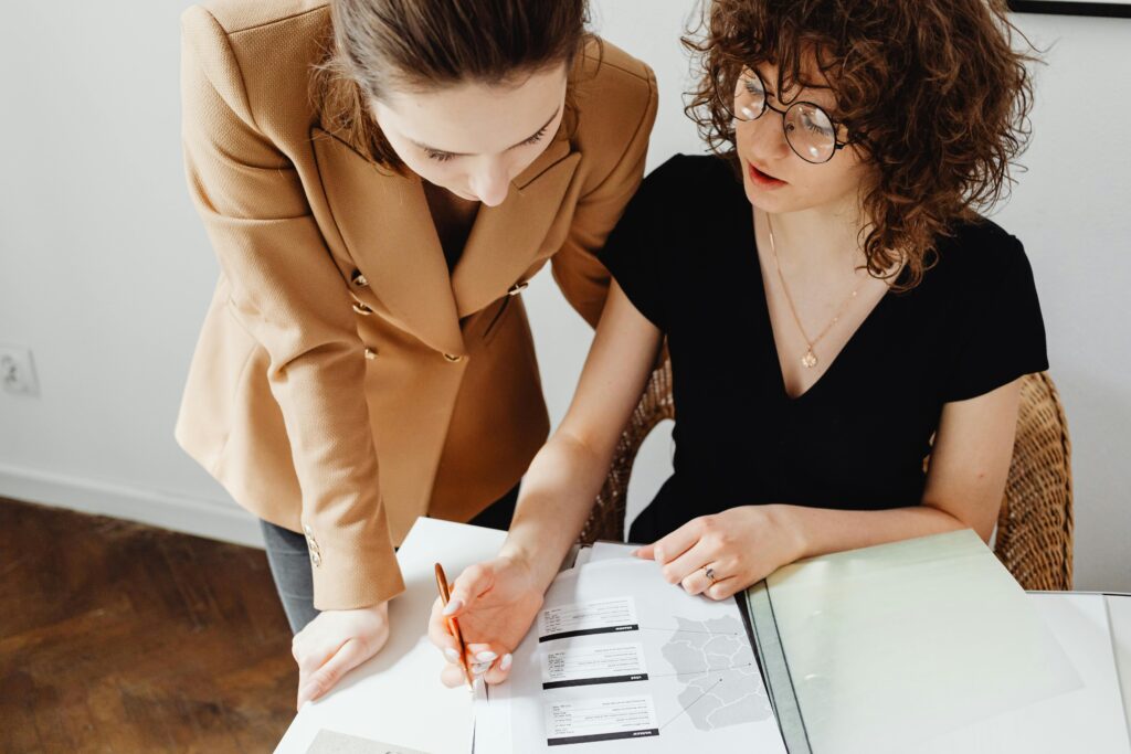 Two businesswomen collaborating over documents, one guiding the other through a decision