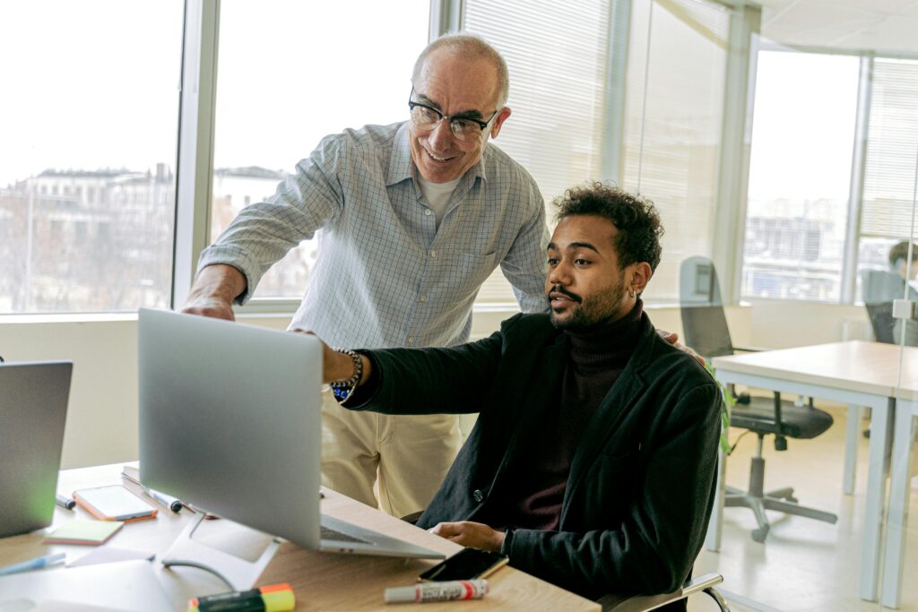 A senior manager leans in to guide a younger colleague at a laptop, embodying servant leadership through mentoring and support